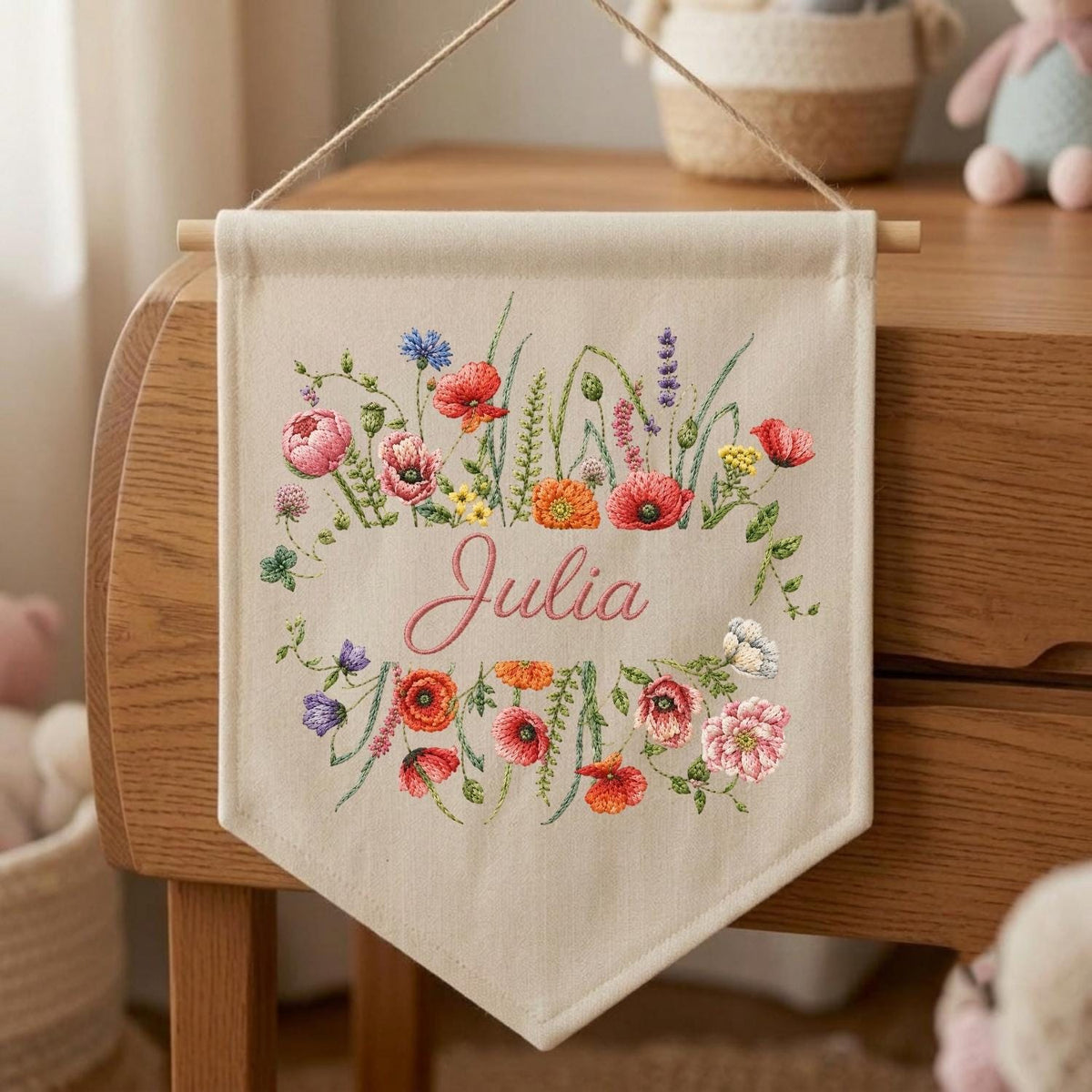 May include: A beige fabric wall banner featuring an embroidered floral wreath with red, pink, orange, and blue wildflowers surrounding the name Julia in pink cursive script. The banner hangs from a wooden dowel with a jute twine string.