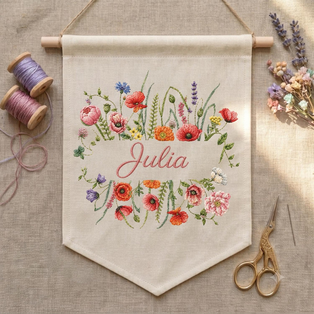 May include: A beige fabric wall banner featuring an embroidered floral wreath in shades of red, pink, orange, and purple surrounding the name Julia in cursive script. The banner hangs from a wooden dowel and is accompanied by two spools of thread.