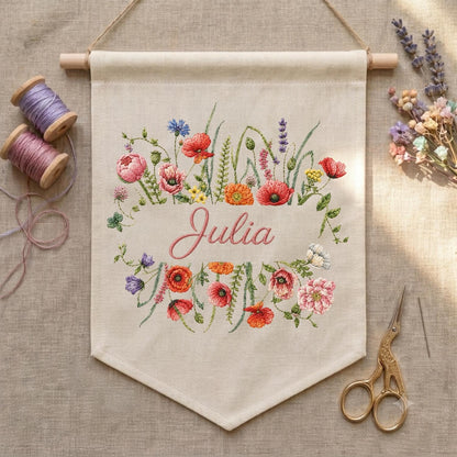 May include: A beige fabric wall banner featuring an embroidered floral wreath in shades of red, pink, orange, and purple surrounding the name Julia in cursive script. The banner hangs from a wooden dowel and is accompanied by two spools of thread.