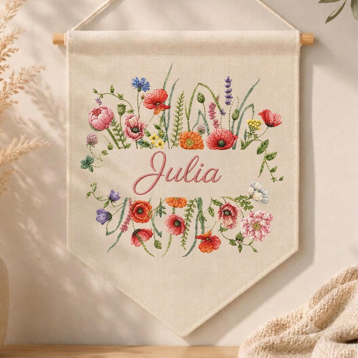 May include: A cream-colored fabric wall hanging featuring an embroidered floral wreath of red, pink, orange, and purple wildflowers surrounding the name Julia in pink cursive script. The banner is suspended from a wooden dowel with a white cord.