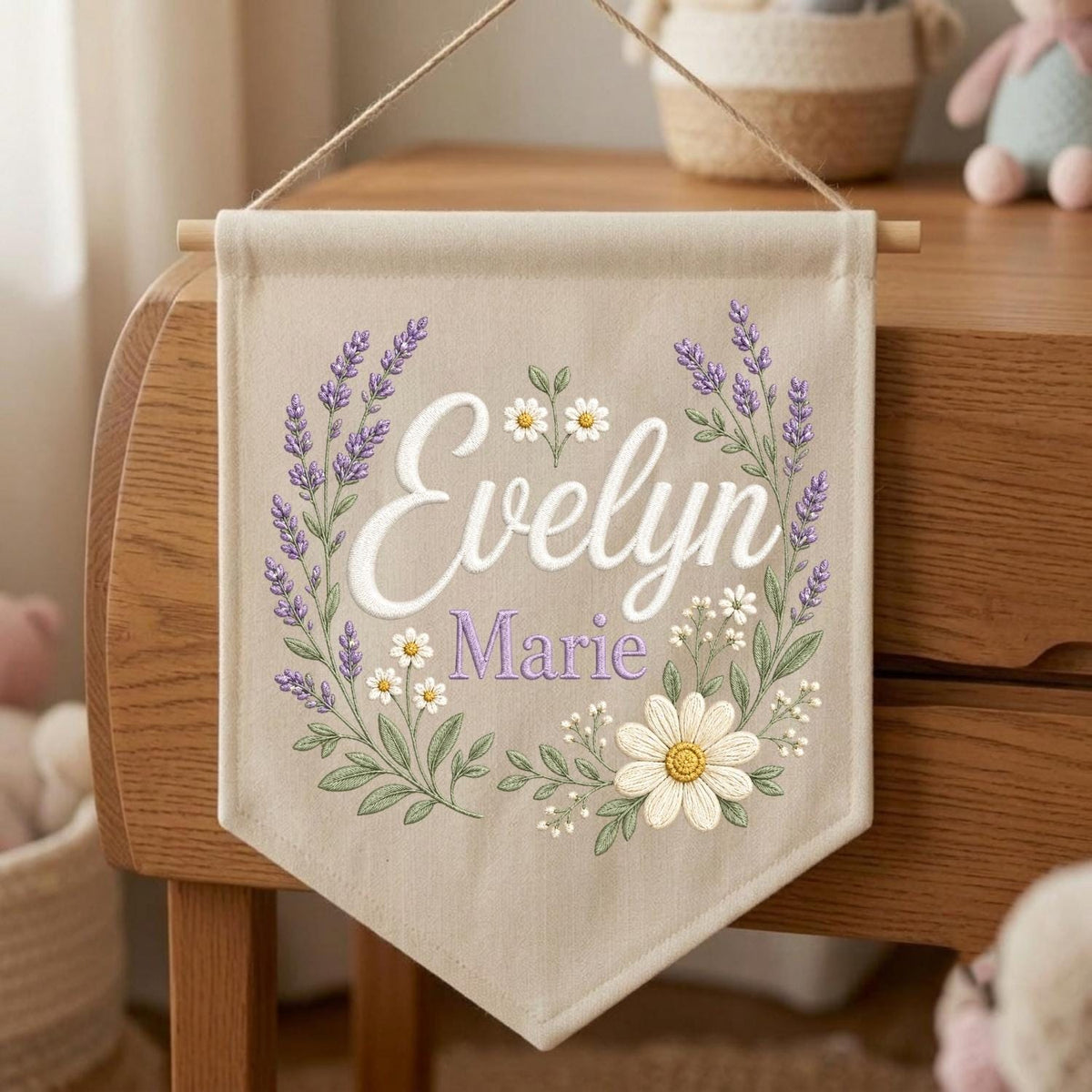 May include: A beige fabric wall banner featuring the embroidered name Evelyn Marie in white and purple script. The text is surrounded by a floral wreath of purple lavender sprigs and white daisies with green stems and leaves, suspended by a string.