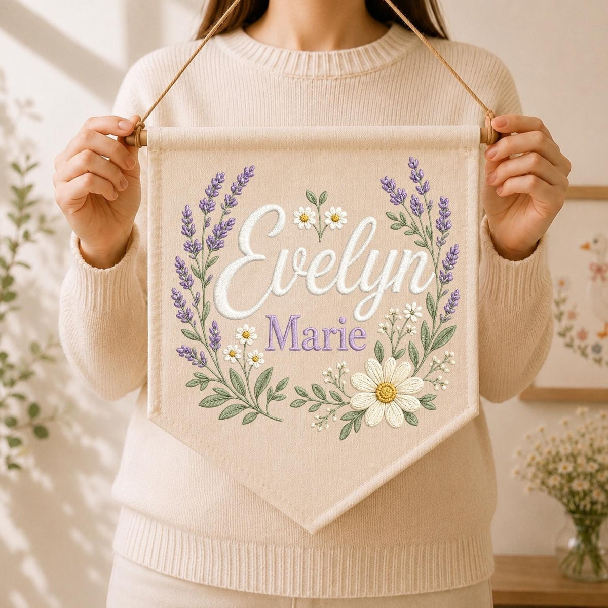 May include: A cream-colored fabric wall banner featuring the embroidered name Evelyn Marie in white and purple script. The text is surrounded by a floral wreath design consisting of purple lavender sprigs and white daisies with green leaves.