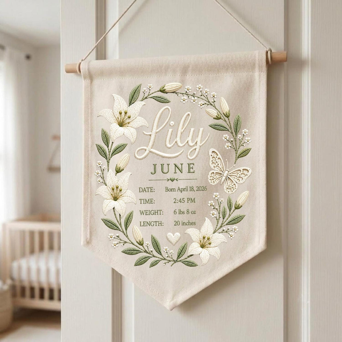 May include: A beige fabric wall banner featuring a floral wreath of white lilies and green leaves surrounding a butterfly. The text reads Lily, June, and birth details: Born April 18, 2026, 2:45 PM, 6 lbs 8 oz, and 20 inches, with a small heart icon.
