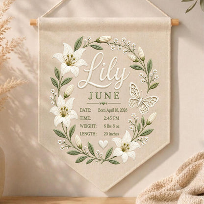 May include: A beige fabric wall hanging featuring a floral wreath of white lilies and green leaves surrounding the name Lily, the month June, and birth details: Born April 18, 2026, 2:45 PM, 6 lbs 8 oz, and 20 inches, with a butterfly illustration.