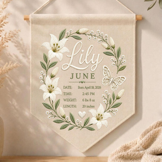 May include: A beige fabric wall hanging featuring a floral wreath of white lilies and green leaves surrounding the name Lily, the month June, and birth details: Born April 18, 2026, 2:45 PM, 6 lbs 8 oz, and 20 inches, with a butterfly illustration.
