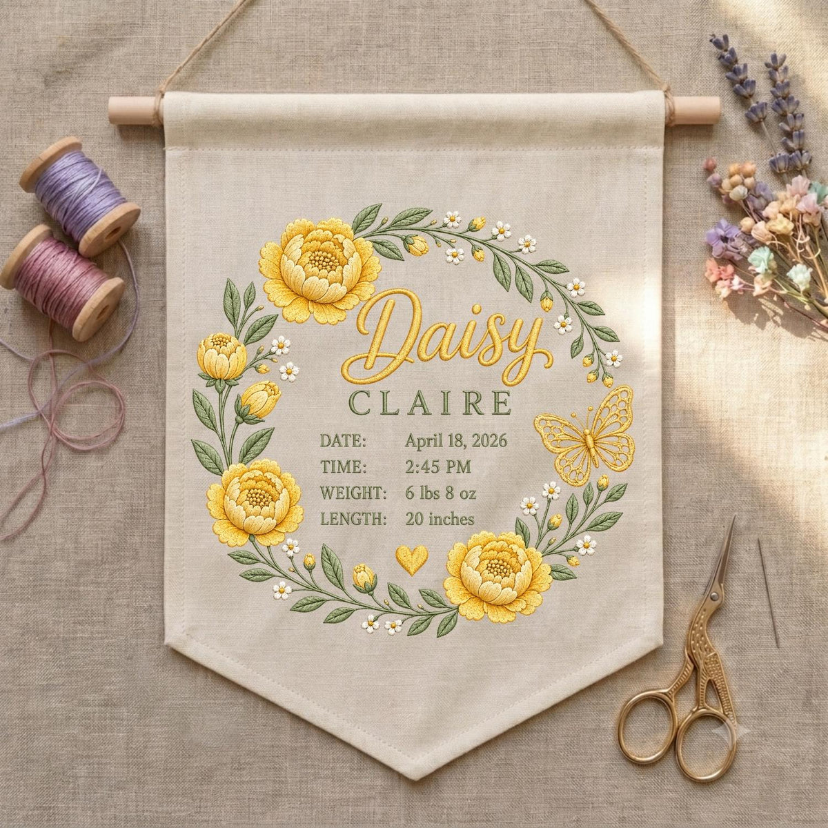 May include: A beige fabric wall hanging featuring an embroidered wreath of yellow flowers and green leaves. The text reads Daisy CLAIRE, DATE: April 18, 2026, TIME: 2:45 PM, WEIGHT: 6 lbs 8 oz, LENGTH: 20 inches, with a small yellow heart and butterfly.