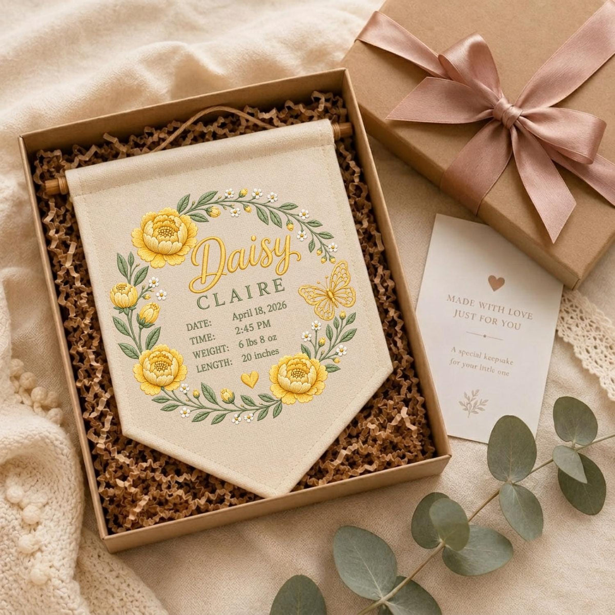 May include: A cream-colored fabric wall banner featuring a yellow floral wreath, a butterfly, and the text Daisy Claire, April 18, 2026, 2:45 PM, 6 lbs 8 oz, and 20 inches. The banner is inside a gift box with a pink ribbon and a note.