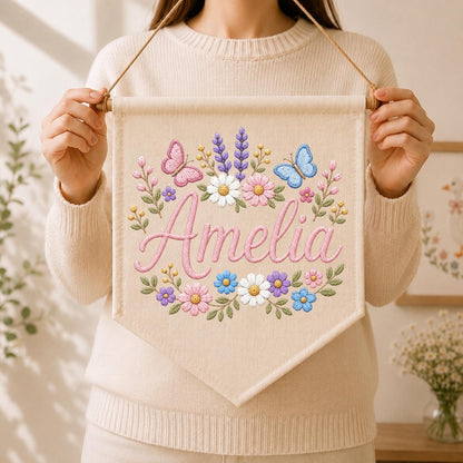 May include: A beige fabric wall banner featuring the embroidered name Amelia in pink script, surrounded by a colorful floral wreath with pink and blue butterflies, white daisies, purple lavender, and green leaves, suspended by a wooden rod and string.
