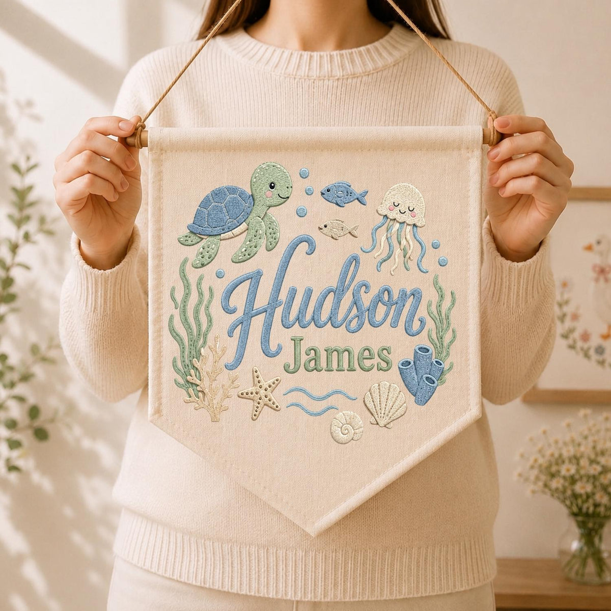 May include: A cream-colored fabric wall banner featuring embroidered ocean life including a green sea turtle, a jellyfish, fish, coral, a starfish, and seashells. The text "Hudson James" is embroidered in blue and green script and block letters.
