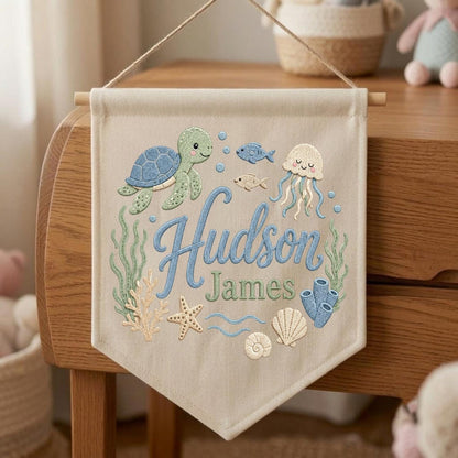 May include: A beige fabric wall banner featuring embroidered ocean life including a green sea turtle, a jellyfish, a starfish, seashells, and coral. The text "Hudson James" is embroidered in blue and green script in the center.