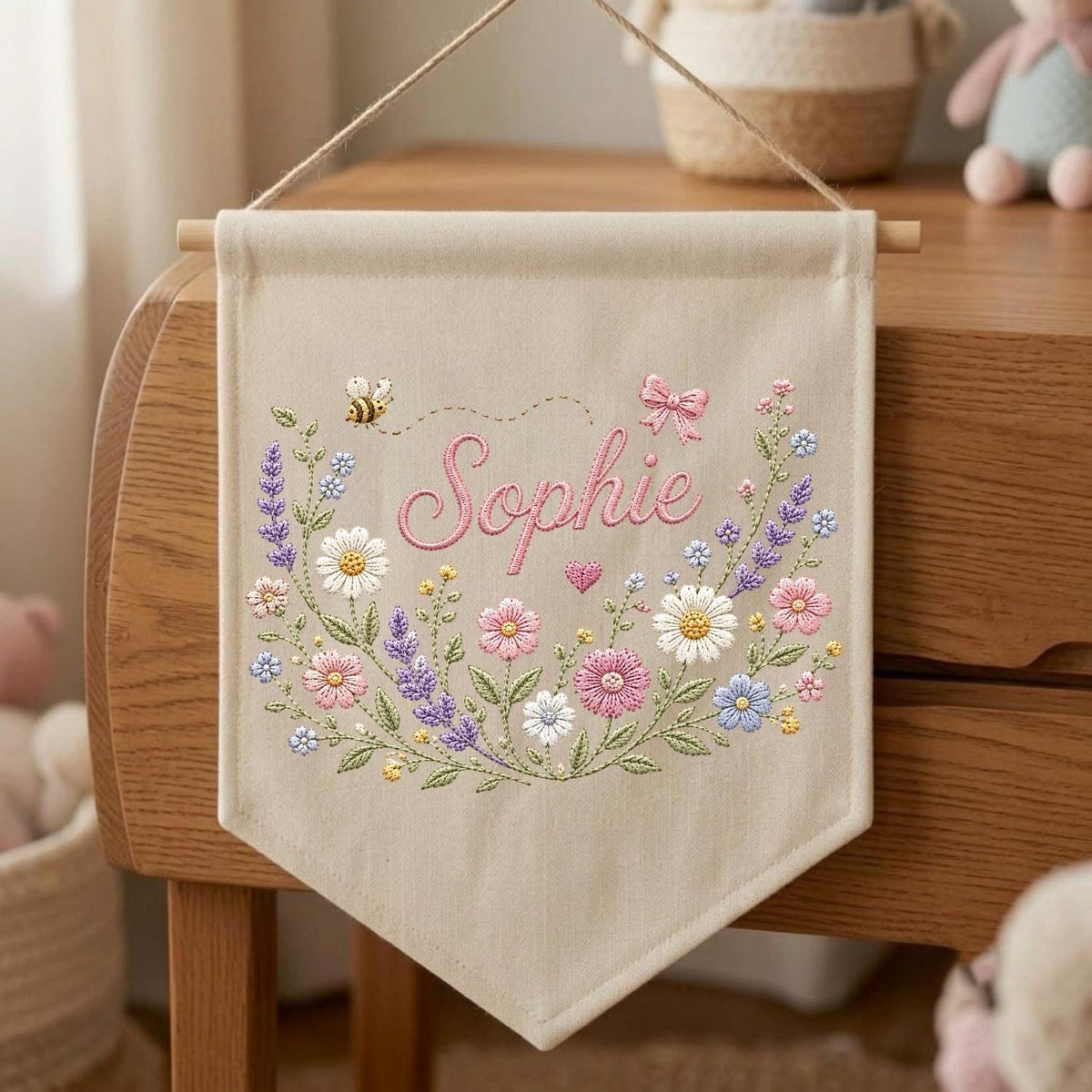 May include: A beige fabric wall banner featuring the embroidered name Sophie in pink script. The design includes a colorful floral wreath with daisies, lavender, and pink flowers, a small bee, a pink bow, and a heart, suspended by a wooden rod and string.