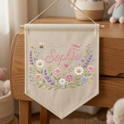 May include: A beige fabric wall banner featuring the embroidered name Sophie in pink script. The design includes a colorful floral wreath with daisies, lavender, and pink flowers, a small bee, a pink bow, and a heart, suspended by a wooden rod and string.