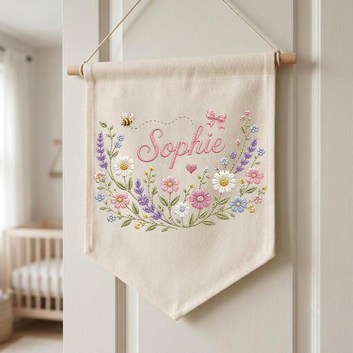May include: A cream-colored fabric wall banner featuring the embroidered name Sophie in pink script. The design includes a colorful floral wreath with daisies, lavender, and pink flowers, a small bee, a pink bow, and a tiny heart, all suspended from a wooden rod.