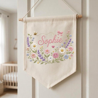 May include: A cream-colored fabric wall banner featuring the embroidered name Sophie in pink script. The design includes a colorful floral wreath with daisies, lavender, and pink flowers, a small bee, a pink bow, and a tiny heart, all suspended from a wooden rod.