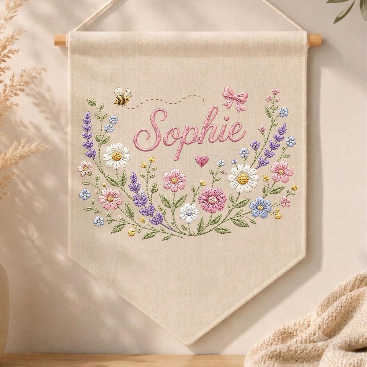 May include: A beige fabric wall banner featuring the embroidered name Sophie in pink script. The design includes a colorful floral wreath with lavender, daisies, and pink flowers, accented by a small bee, a pink bow, and a heart.