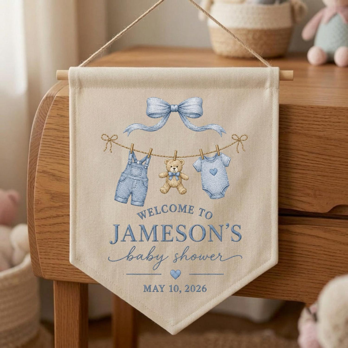 May include: A beige fabric wall banner featuring a blue bow, a clothesline with blue overalls, a teddy bear, and a blue onesie. The text reads: WELCOME TO JAMESON'S baby shower MAY 10, 2026. The banner hangs from a wooden dowel with a string.