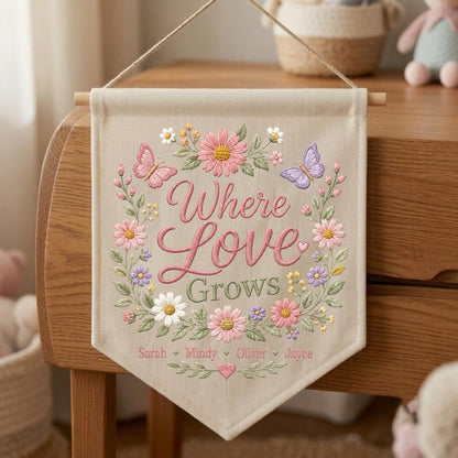 May include: A beige fabric wall banner featuring the embroidered text Where Love Grows in pink script. The text is surrounded by a floral wreath with pink, white, and purple flowers, green leaves, and two butterflies. Four names are embroidered below.