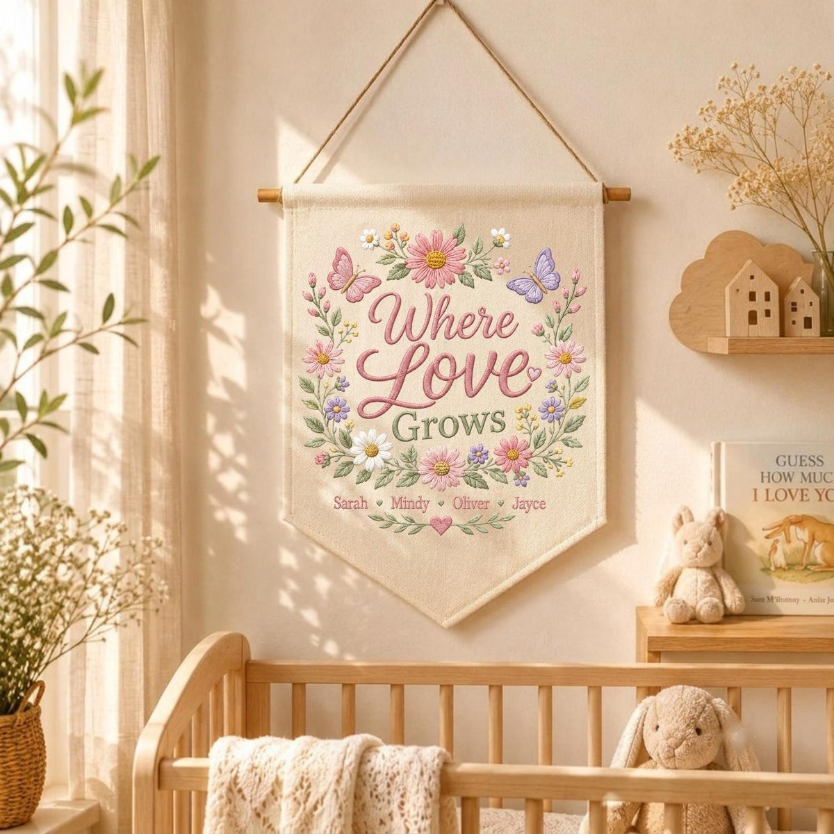 May include: A cream-colored fabric wall hanging featuring an embroidered floral wreath with butterflies and the text Where Love Grows. Below the text, the names Sarah, Mindy, Oliver, and Jayce are embroidered in a script font with small hearts.