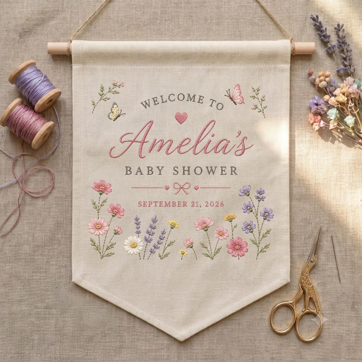 May include: A beige fabric wall banner featuring embroidered floral motifs, butterflies, and the text Welcome to Amelia's Baby Shower September 21, 2026. The banner hangs from a wooden rod and is accompanied by two spools of thread and scissors.