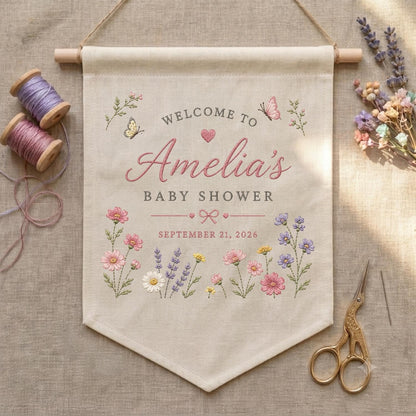 May include: A beige fabric wall banner featuring embroidered floral motifs, butterflies, and the text Welcome to Amelia's Baby Shower September 21, 2026. The banner hangs from a wooden rod and is accompanied by two spools of thread and scissors.