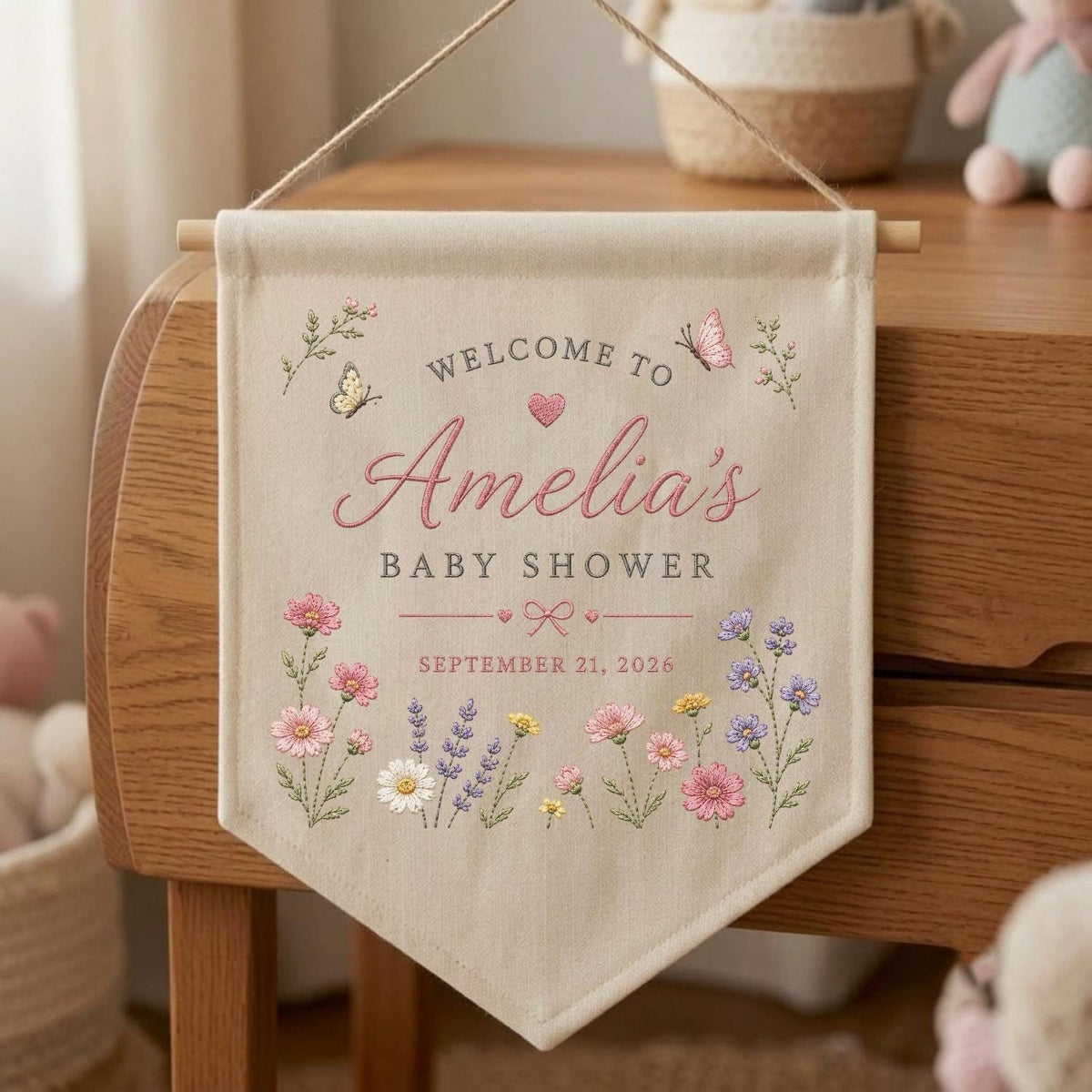 May include: A beige fabric wall banner featuring the text "WELCOME TO Amelia's BABY SHOWER" and "SEPTEMBER 21, 2026". The design includes embroidered pink, purple, and white wildflowers, a small heart, a bow, and two butterflies.