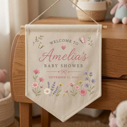 May include: A beige fabric wall banner featuring the text "WELCOME TO Amelia's BABY SHOWER" and "SEPTEMBER 21, 2026". The design includes embroidered pink, purple, and white wildflowers, a small heart, a bow, and two butterflies.