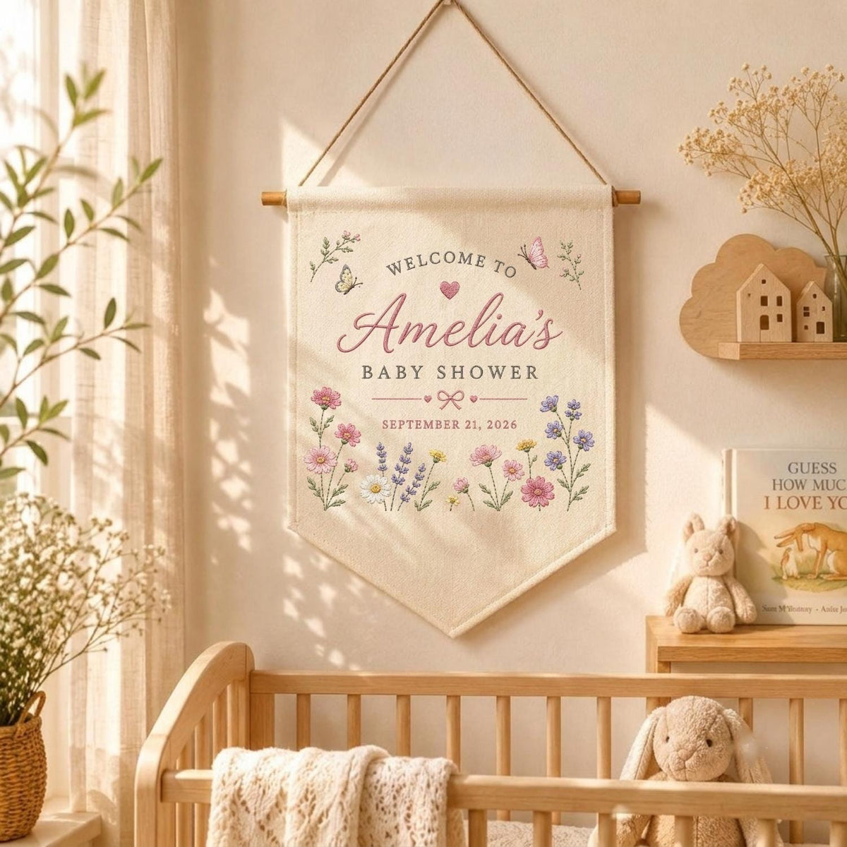 May include: A cream-colored fabric wall banner hanging from a wooden rod with a string. The banner features the text "WELCOME TO Amelia's BABY SHOWER" and "SEPTEMBER 21, 2026" in pink and gray, decorated with colorful floral illustrations and butterflies.