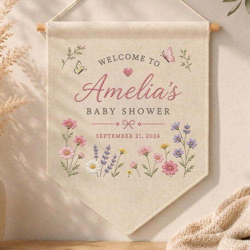May include: A cream-colored fabric wall banner featuring the text "WELCOME TO Amelia's BABY SHOWER" and "SEPTEMBER 21, 2026". The design includes embroidered pink and purple wildflowers, two butterflies, a small pink heart, and a bow graphic.