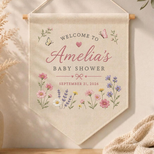 May include: A cream-colored fabric wall banner featuring the text "WELCOME TO Amelia's BABY SHOWER" and "SEPTEMBER 21, 2026". The design includes embroidered pink and purple wildflowers, two butterflies, a small pink heart, and a bow graphic.