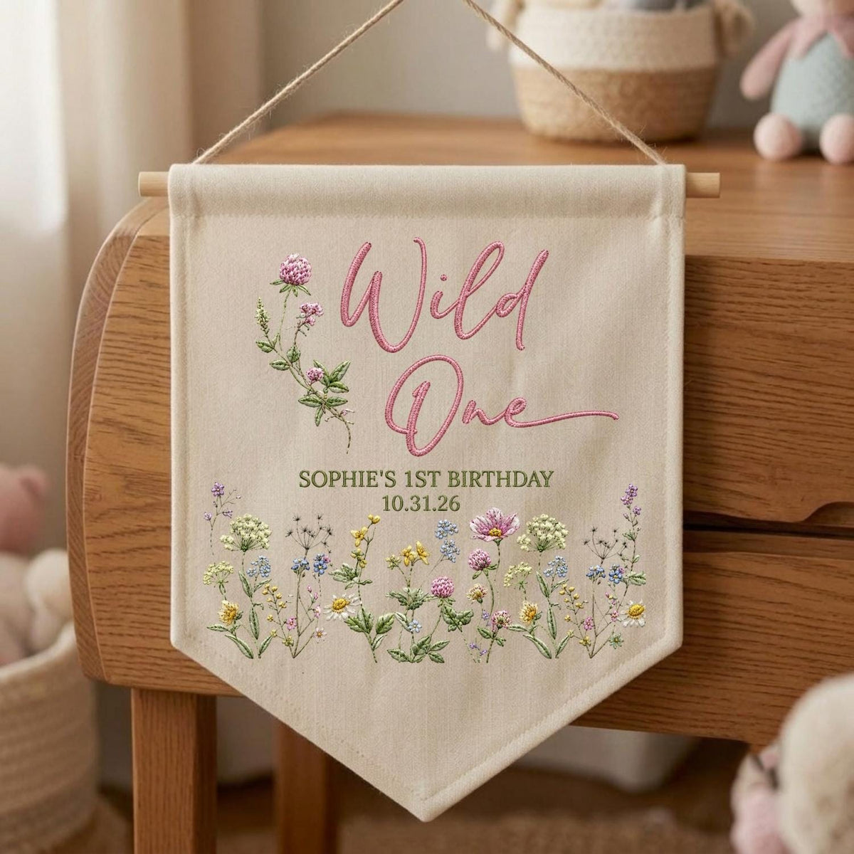 May include: A cream-colored fabric wall banner featuring the pink embroidered text Wild One, with the printed text SOPHIE'S 1ST BIRTHDAY 10.31.26. The banner is decorated with colorful illustrations of wildflowers and clover stems.