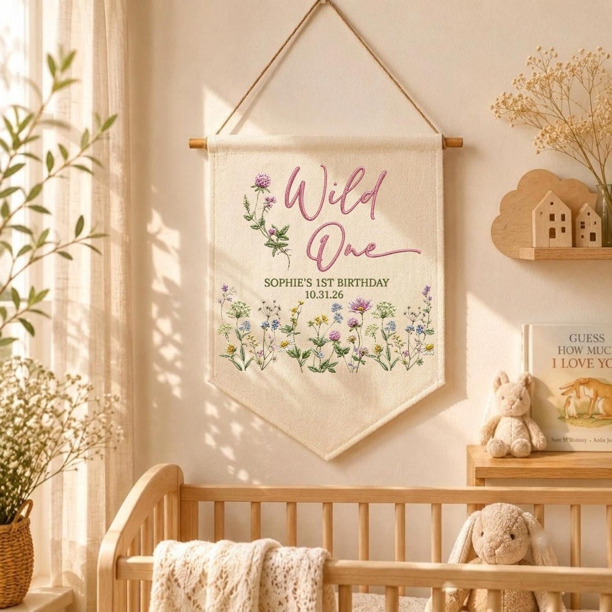 May include: A cream-colored fabric wall banner hanging from a wooden rod with a string. The banner features the pink text Wild One, the words SOPHIE'S 1ST BIRTHDAY 10.31.26, and a colorful illustration of various wildflowers across the bottom section.