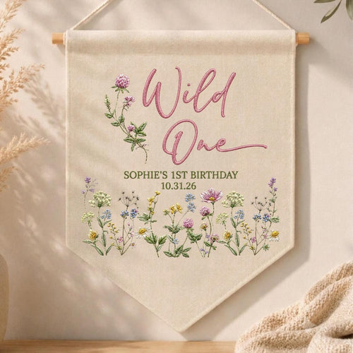 May include: A cream-colored fabric wall banner hanging from a wooden dowel. The text reads Wild One, SOPHIE'S 1ST BIRTHDAY, and 10.31.26. The design features embroidered illustrations of pink clover and various colorful wildflowers at the bottom.