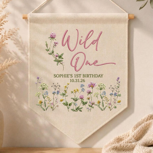 May include: A cream-colored fabric wall banner hanging from a wooden dowel. The text reads Wild One, SOPHIE'S 1ST BIRTHDAY, and 10.31.26. The design features embroidered illustrations of pink clover and various colorful wildflowers at the bottom.