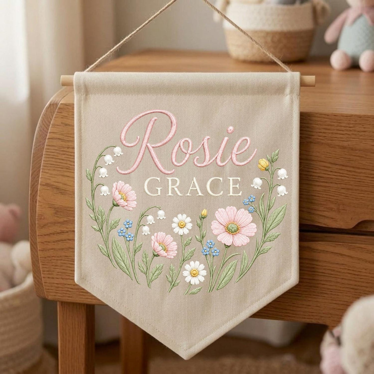 May include: A beige fabric wall banner featuring the name Rosie Grace in pink and white script and block lettering. The design includes a floral wreath illustration with pink cosmos, white daisies, blue forget-me-nots, and lily of the valley.