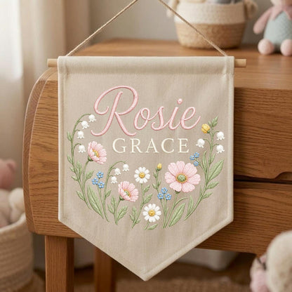 May include: A beige fabric wall banner featuring the name Rosie Grace in pink and white script and block lettering. The design includes a floral wreath illustration with pink cosmos, white daisies, blue forget-me-nots, and lily of the valley.