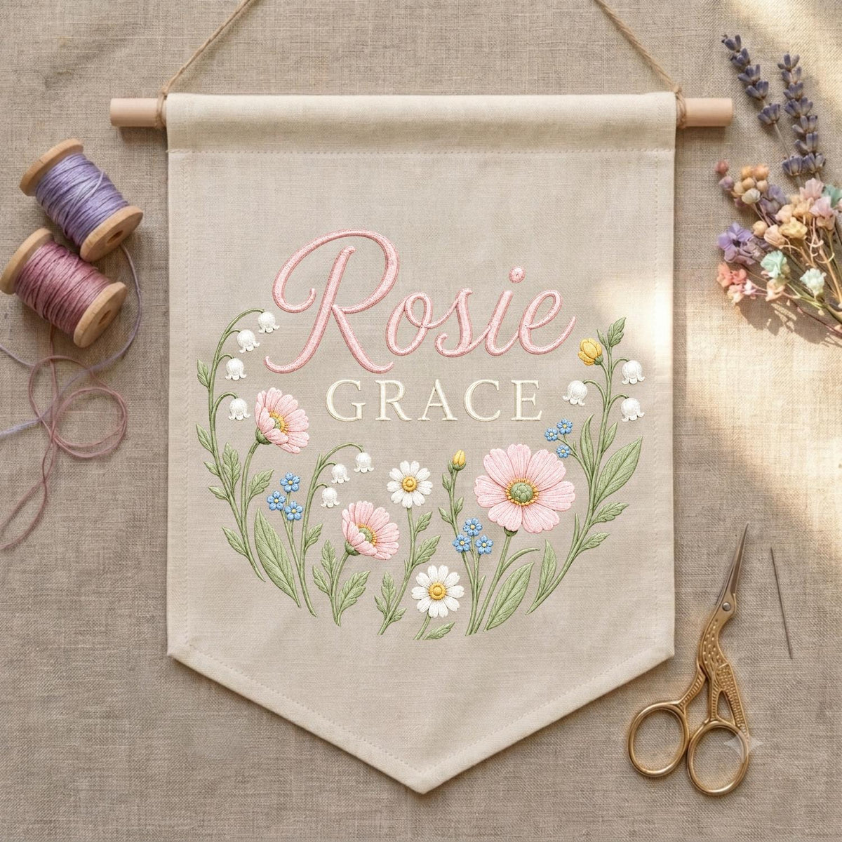 May include: A beige fabric wall banner featuring the embroidered name Rosie Grace in pink script and block lettering. The design includes a floral wreath of pink, white, and blue flowers with green stems and leaves, suspended from a wooden rod.