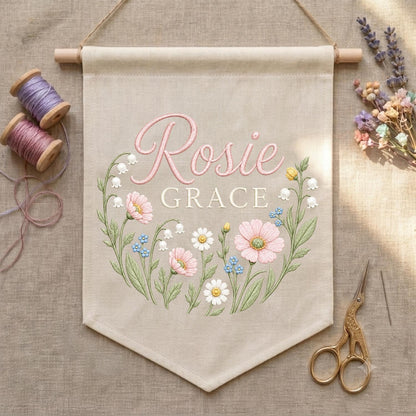 May include: A beige fabric wall banner featuring the embroidered name Rosie Grace in pink script and block lettering. The design includes a floral wreath of pink, white, and blue flowers with green stems and leaves, suspended from a wooden rod.