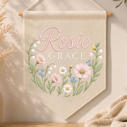 May include: A beige fabric wall banner featuring the name Rosie Grace in pink and white lettering. The design includes a floral wreath illustration with pink cosmos, white daisies, blue forget-me-nots, and lily of the valley stems.