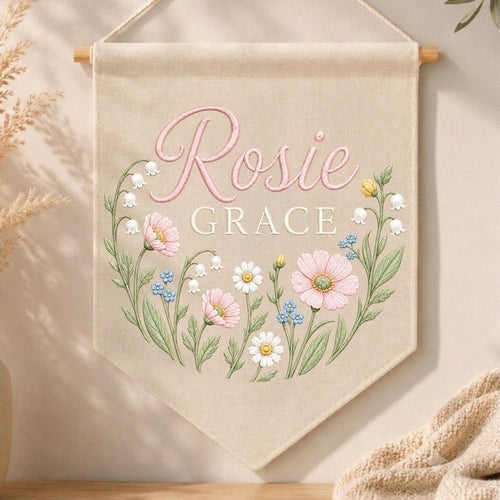 May include: A beige fabric wall banner featuring the name Rosie Grace in pink and white lettering. The design includes a floral wreath illustration with pink cosmos, white daisies, blue forget-me-nots, and lily of the valley stems.