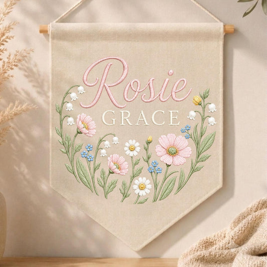 May include: A beige fabric wall banner featuring the name Rosie Grace in pink and white lettering. The design includes a floral wreath illustration with pink cosmos, white daisies, blue forget-me-nots, and lily of the valley stems.