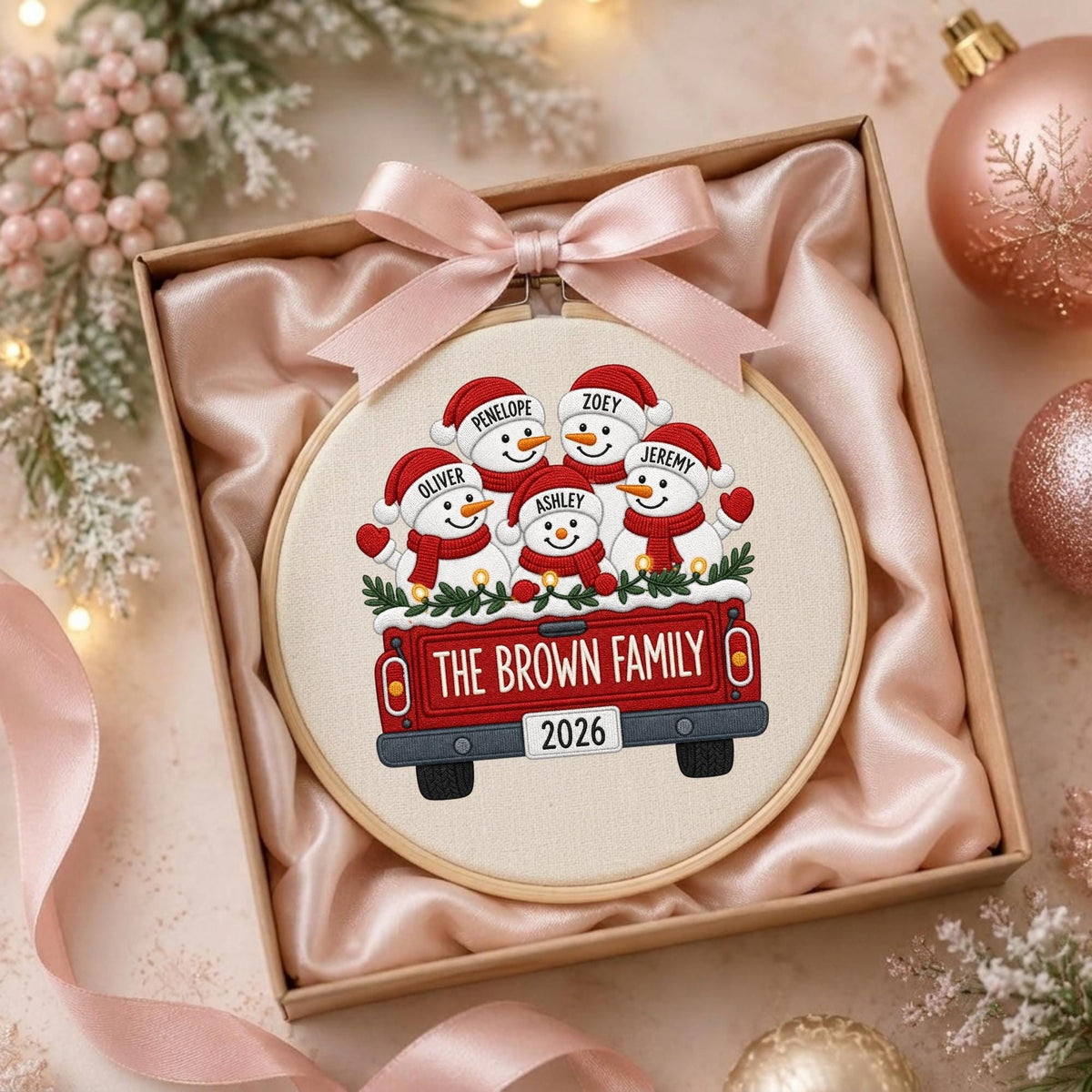 May include: A round embroidery hoop containing a fabric panel with a printed illustration of five snowmen in a red pickup truck. The truck features the text THE BROWN FAMILY and 2026. The snowmen are labeled Penelope, Zoey, Oliver, Ashley, and Jeremy.