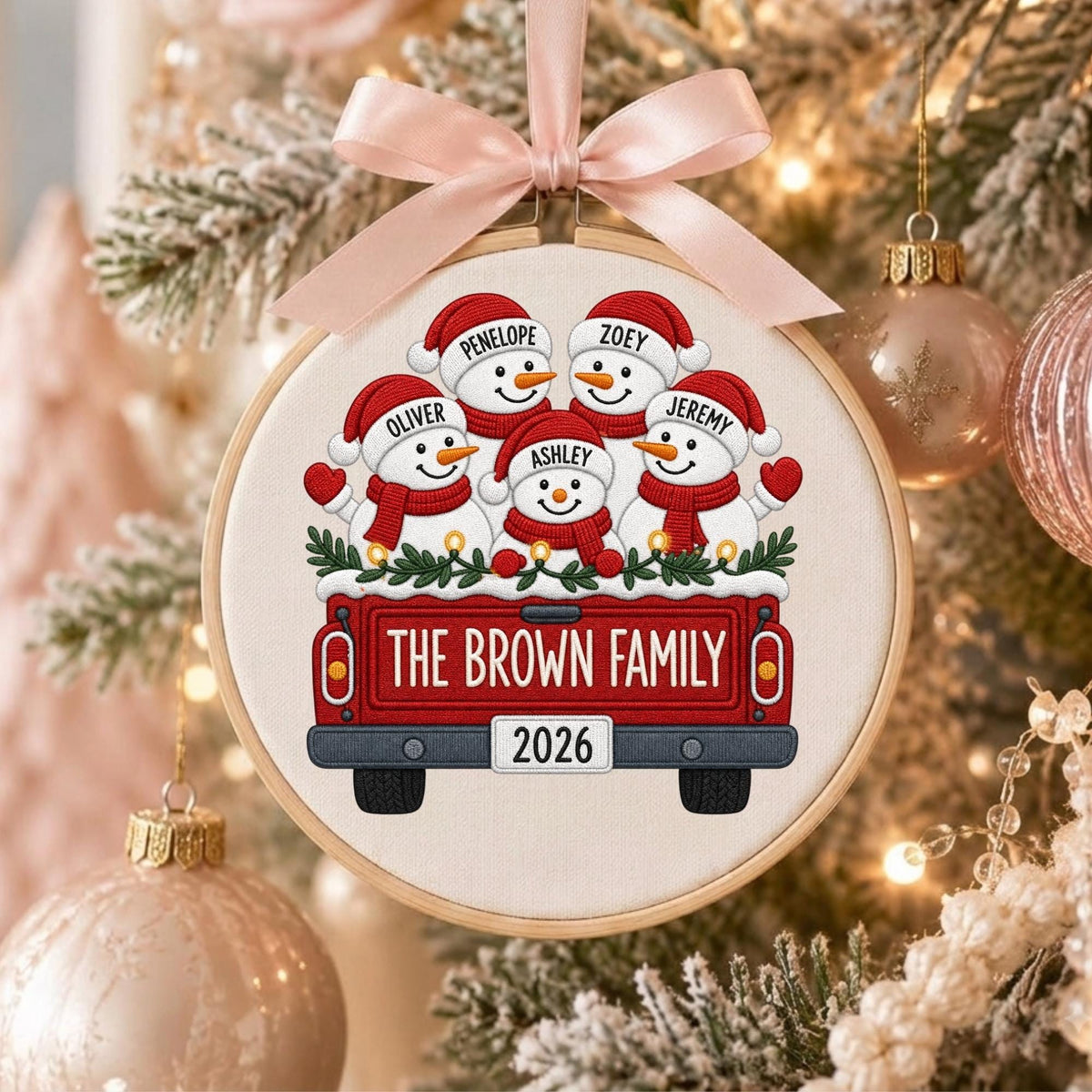 May include: A round wooden embroidery hoop ornament featuring a light fabric background with a red truck illustration. The truck carries five snowmen wearing red hats and scarves, labeled Penelope, Zoey, Oliver, Ashley, and Jeremy. Text reads THE BROWN FAMILY and 2026. A light pink ribbon bow is attached to the top.