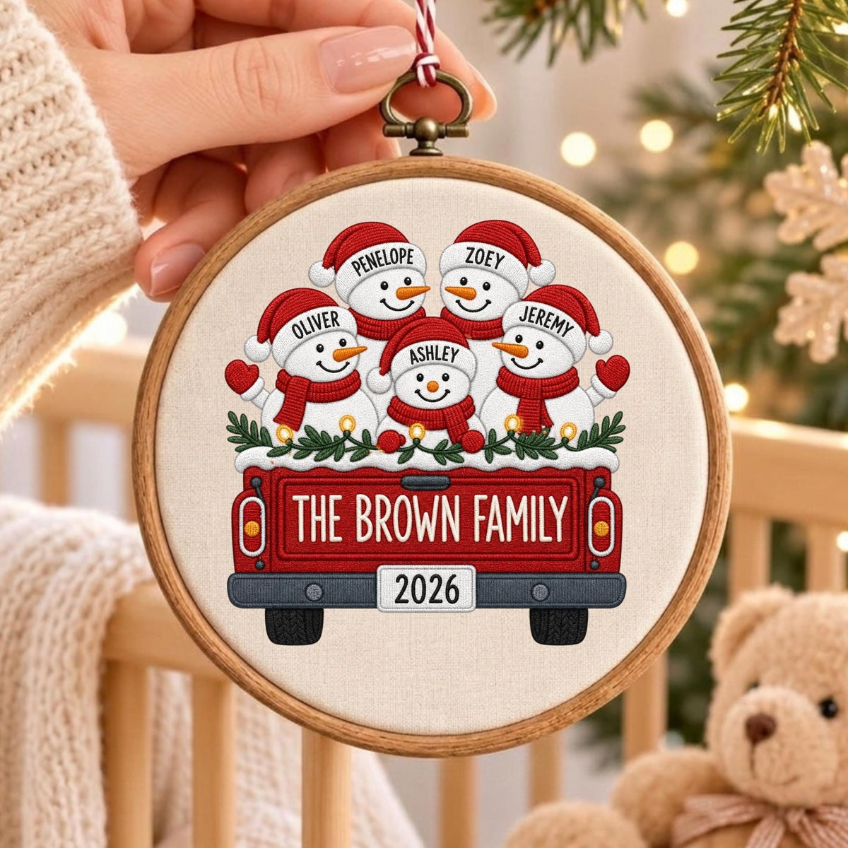 May include: A round wooden embroidery hoop ornament featuring a fabric insert with a red truck illustration. Five snowmen wearing red hats and scarves are in the truck bed. Text reads THE BROWN FAMILY and 2026, with names Penelope, Zoey, Oliver, Jeremy, and Ashley.