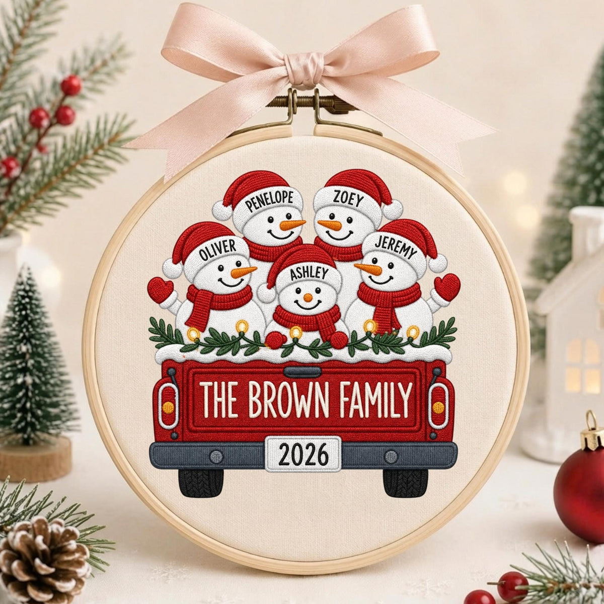May include: A circular embroidery hoop featuring a fabric panel with a red truck illustration carrying five snowmen wearing red hats and scarves. The snowmen are labeled Penelope, Zoey, Oliver, Jeremy, and Ashley. The truck reads THE BROWN FAMILY 2026.