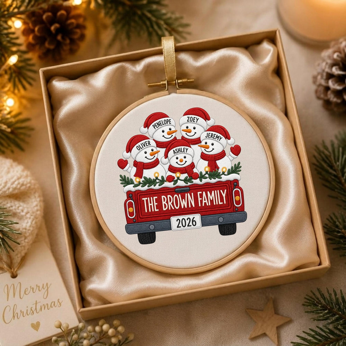 May include: A round embroidery hoop ornament featuring a printed illustration of five snowmen wearing red hats and scarves inside a red pickup truck. The truck bed displays the text THE BROWN FAMILY and a license plate with the year 2026.
