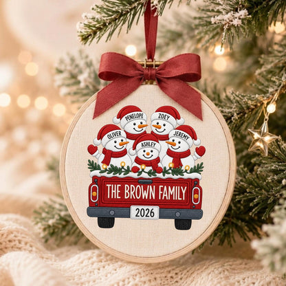 May include: A round wooden embroidery hoop ornament featuring a fabric insert with a printed illustration of five snowmen in a red pickup truck. The truck bed displays the text THE BROWN FAMILY and 2026. A dark red ribbon bow is tied at the top.