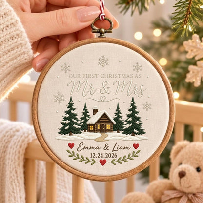 May include: A round wooden embroidery hoop ornament featuring a cream-colored fabric center with a printed winter scene of a cabin, pine trees, and snowflakes. Text reads: OUR FIRST CHRISTMAS AS Mr &amp; Mrs, Emma &amp; Liam, and 12.24.2026.