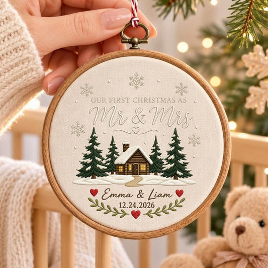 May include: A round wooden embroidery hoop ornament featuring a cream-colored fabric center with a printed winter scene of a cabin, pine trees, and snowflakes. Text reads: OUR FIRST CHRISTMAS AS Mr &amp; Mrs, Emma &amp; Liam, and 12.24.2026.