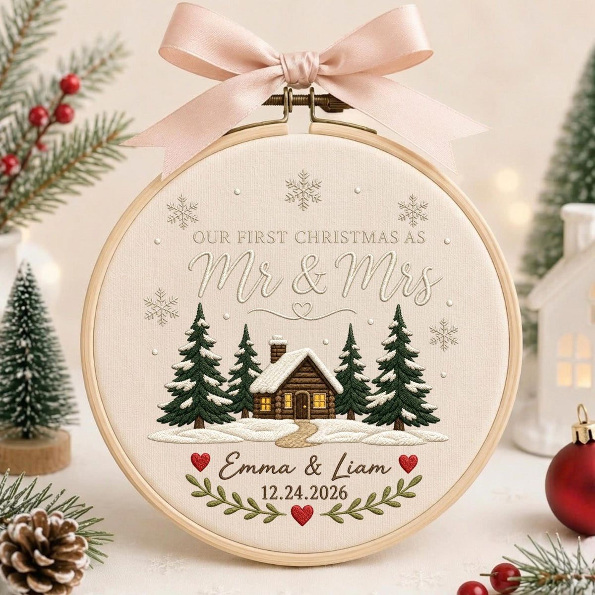 May include: A circular embroidery hoop featuring cream fabric with a stitched winter scene of a log cabin, pine trees, and snowflakes. Text reads: Our First Christmas as Mr &amp; Mrs, Emma &amp; Liam, 12.24.2026. A light pink ribbon is tied at the top.
