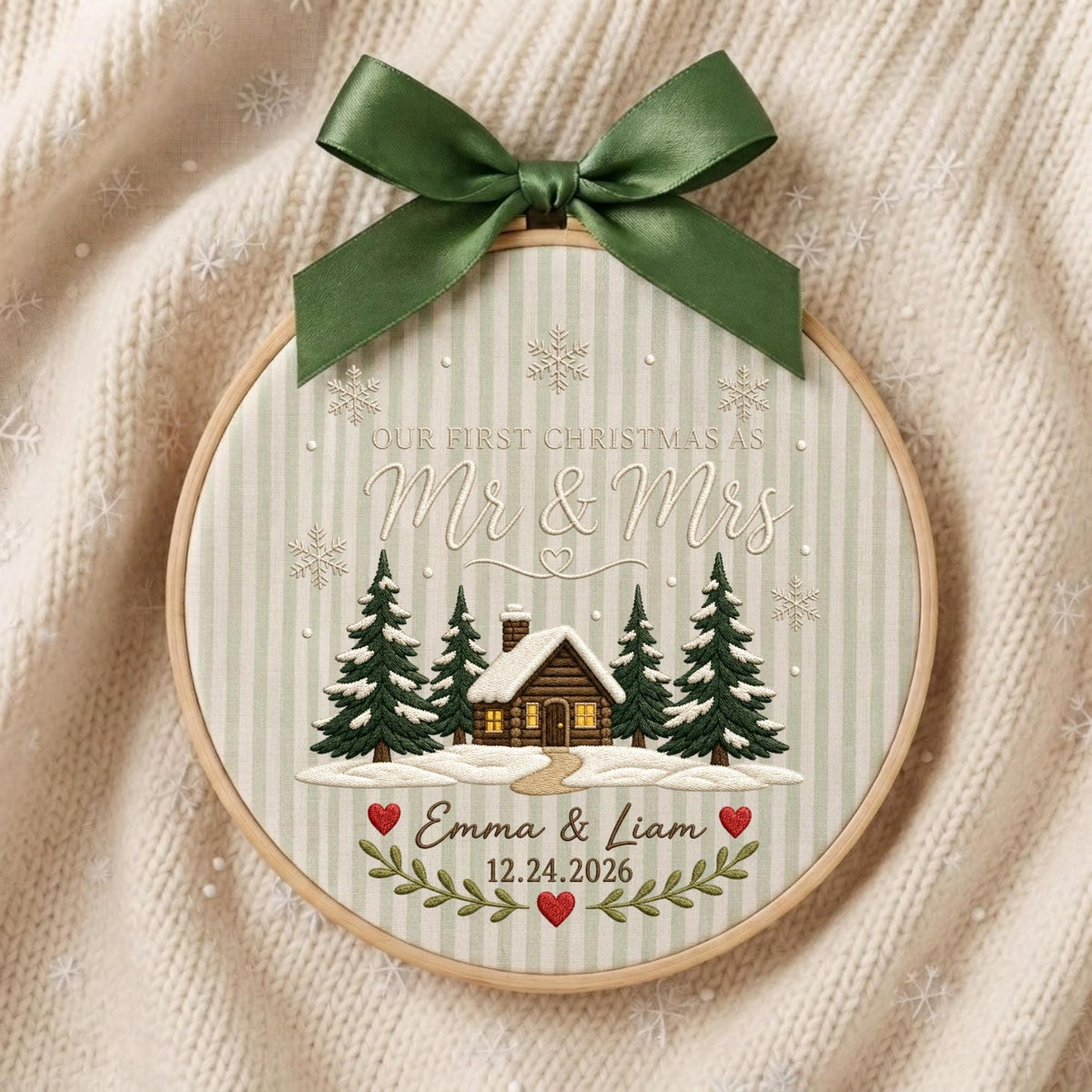 May include: A round embroidery hoop ornament featuring a green satin bow at the top. The fabric has a light green striped pattern with embroidered snowflakes, a cabin in a snowy forest, and the text "OUR FIRST CHRISTMAS AS Mr &amp; Mrs Emma &amp; Liam 12.24.2026" with red hearts.